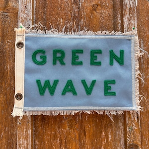 May include: A rectangular light blue fabric flag with frayed edges and a white canvas header featuring two metal grommets. The words GREEN WAVE are stitched onto the center of the flag in bold, dark green felt lettering with a visible texture.
