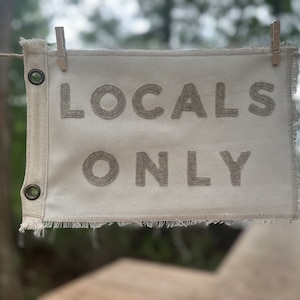 May include: A white canvas sign with frayed edges hangs from a rope with clothespins. The sign reads "LOCALS ONLY" in large, beige, felt letters.