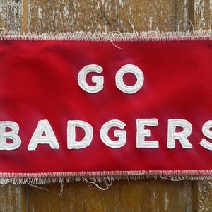 Op de afbeelding: Een rode vlag met de woorden "GO BADGERS" in witte, gestikte letters. De vlag heeft een gerafelde rand en grommets aan de linkerkant. De vlag is rechthoekig en heeft een witte rand.