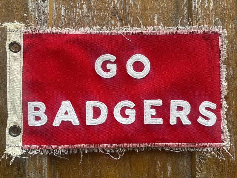 Op de afbeelding: Een rode vlag met de woorden "GO BADGERS" in witte, gestikte letters. De vlag heeft een gerafelde rand en grommets aan de linkerkant. De vlag is rechthoekig en heeft een witte rand.