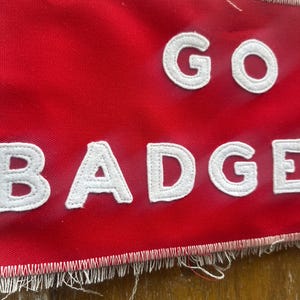 Op de afbeelding: Een rode banner met de woorden "GO BADGERS" in witte, gestikte letters. De banner heeft een gerafelde rand. De letters zijn vetgedrukt en zonder schreef. De banner is gemaakt van rode stof.