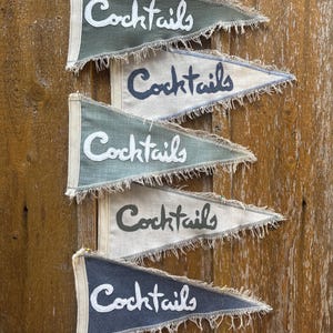 Peut inclure: Cinq fanions avec le mot "Cocktails" écrit dessus en différentes couleurs et styles. Les fanions sont accrochés à un mur en bois.