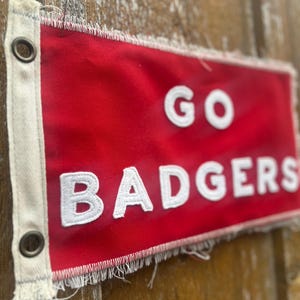 Op de afbeelding: Een rode vlag met de woorden "GO BADGERS" in witte, gestikte letters. De vlag heeft een gerafelde rand en ogen om op te hangen. De achtergrond is een houten oppervlak.