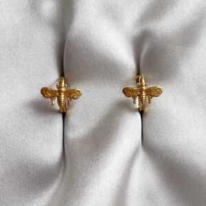 May include: Two gold-coloured bee-shaped hoop earrings. The earrings are presented on a white surface. The bees have detailed wings and bodies, and the hoops are small and circular.