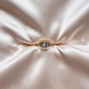 May include: A gold ring featuring three rectangular, iridescent gray stones arranged horizontally. The center stone is larger than the two flanking stones, all set in a simple gold band that encircles the finger with a polished, metallic finish.