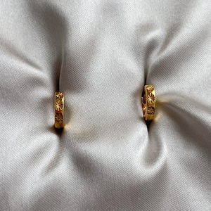 May include: A pair of gold hoop earrings. Each earring has star-shaped cutouts along its surface. The earrings are presented on a white, silky fabric, contrasting with the shiny gold. The earrings are approximately 1.5 cm in diameter.