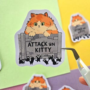 Op de afbeelding: Een sticker met een oranje en witte pluizige kat die over een grijze stenen muur kijkt met de tekst "ATTACK N KITTY". Kleine zwarte kat silhouetten staan aan de basis. Andere stickers zijn ook zichtbaar.