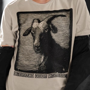 May include: A person wearing a light brown t-shirt with a black and white illustration of a goat's head. The text "EMBRACE YOUR SHADOW" is printed below the image.