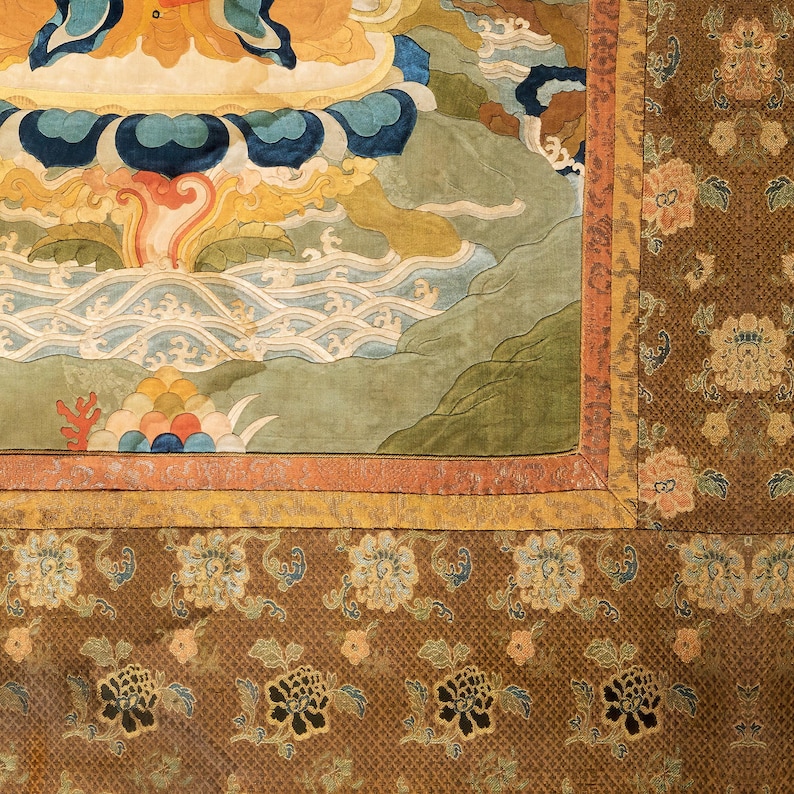 May include: Close-up of a section of a traditional tapestry. The tapestry features a detailed design with a color palette of blues, greens, oranges, and browns. The border has a repeating floral pattern. The central design depicts a landscape with stylized clouds and water.
