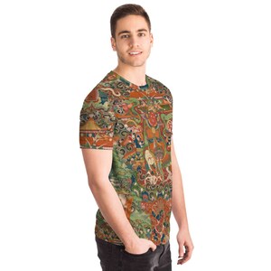 May include: A short-sleeved t-shirt featuring a detailed, colorful illustration. The design incorporates various figures, buildings, and natural elements in shades of orange, green, blue, and red. The shirt is worn by a person.