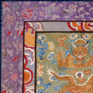 Pode incluir: Close-up de um drag&atilde;o bordado a ouro sobre um fundo verde. O drag&atilde;o &eacute; rodeado por nuvens azuis e amarelas. A borda do tecido &eacute; um padr&atilde;o roxo e azul.