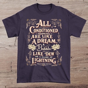 Puede incluir: Una camiseta de color morado oscuro con un diseño gráfico de estilo vintage. El diseño presenta un borde floral y el texto "All Conditioned Phenomena Are Like A Dream, A Bubble, Like Dew Or A Flash Of Lightning - Diamond Sutra".