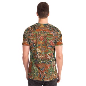May include: A short-sleeved t-shirt featuring a detailed, colorful illustration. The artwork depicts a complex scene with figures, buildings, and intricate patterns in shades of red, green, orange, and blue. The shirt's design covers the entire surface.