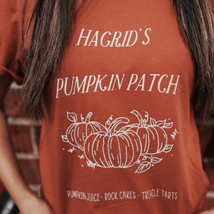 May include: A burnt orange t-shirt with white text that reads "Hagrid's Pumpkin Patch" and a drawing of pumpkins. The text below the pumpkins reads "Pumpkin Juice - Rock Cakes - Treacle Tarts".