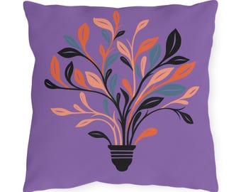 Pode incluir: Almofada quadrada com fundo roxo. A almofada apresenta uma ilustração estilizada de uma planta com folhas pretas, laranja, pêssego e verde-azulado emergindo de uma base preta. O design é centrado na almofada.