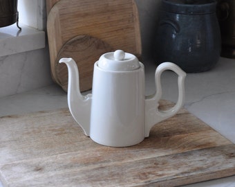May include: A white ceramic teapot with a lid sits on a wooden chopping board. The teapot has a rounded body and a spout on one side. The chopping board is brown and has a natural wood grain.