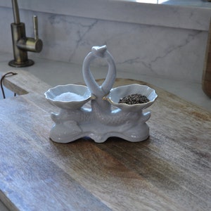 Peut inclure: Salière et poivrière en céramique blanche en forme de cygne, avec une bordure dorée, posés sur une planche à découper en bois.