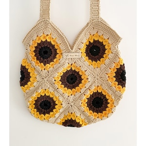 May include: A crocheted tote bag with a sunflower design. The bag is made with beige, brown, yellow, and black yarn. The bag has two straps and a small tag that says "Hand Made".