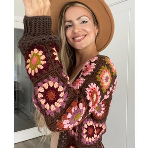 Peut inclure: Un cardigan en crochet marron avec un motif de carrés de grand-mère colorés. Le cardigan a une longue ouverture sur le devant et des manches longues. Le modèle porte un chapeau marron.