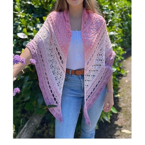 May include: A pink, white, and purple crochet shawl with a lacy pattern. The shawl is draped over a person's shoulders and is worn with a white shirt and blue jeans.