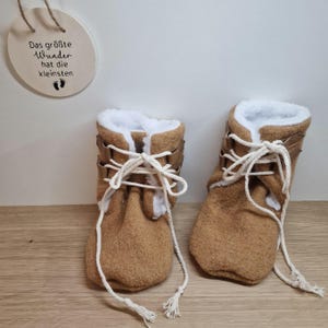 Könnte beinhalten: Ein Paar braune Babyschuhe mit weißem Kunstfellfutter und weißen Schnürsenkeln. Die Schuhe haben ein goldenes Schnallen-Detail und der Text "Das größte Wunder hat die kleinsten" ist im Hintergrund sichtbar.