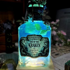 Könnte beinhalten: Dekorative Glasflasche mit leuchtend blauer Flüssigkeit und dem Etikett "Ink of the Kraken". Die Flasche ist mit maritimen Verzierungen und dem Text "Drawn from the Abyss Below" versehen.