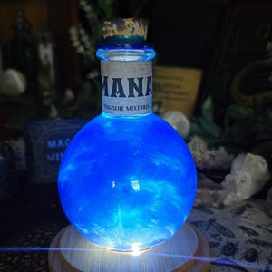 Peut inclure: Une bouteille de potion bleue lumineuse avec le mot "MANA" sur une étiquette. La bouteille est ronde et repose sur une base en bois. Le texte "Magische Mixturen" est également visible. La bouteille est éclairée de l'intérieur.