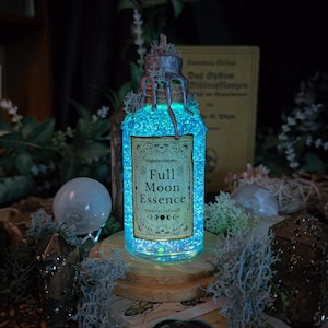 Könnte beinhalten: Eine leuchtende Glasflasche mit der Aufschrift "Full Moon Essence" und einer teal-farbenen Flüssigkeit. Die Flasche ist mit Moos verziert und steht auf einem Holzsockel. Das Design ist mystisch und bezaubernd.