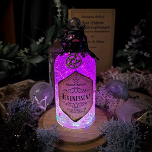 Könnte beinhalten: Eine Glasflasche mit einer lila Flüssigkeit und Glitzer darin. Die Flasche hat einen schwarzen Korkverschluss und ein Etikett mit der Aufschrift "Magische Mixturen Hexenessenz Deine Essenz for Starke Zauber".