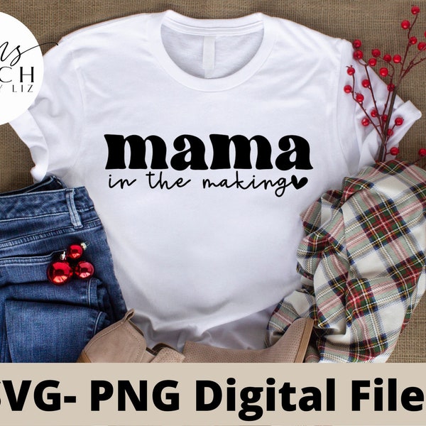 Könnte beinhalten: Weißes T-Shirt mit schwarzem Text, der "mama in the making" mit einem kleinen Herz lautet. Das Shirt liegt auf einer braunen Oberfläche mit einer blauen Jeans, einem karierten Schal und roten Beeren.
