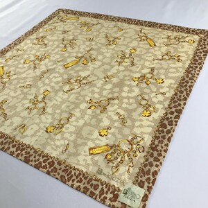Könnte beinhalten: Ein beiger Seidenschal mit braunem Leopardenmuster am Rand. Der Schal zeigt ein sich wiederholendes Muster aus goldenen Ketten und Anhängern, darunter Herzen, Schlüssel und ein Schild mit der Aufschrift "Krizia".