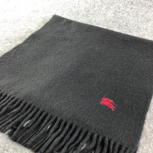 May include: A black scarf with a fringed edge and a small red emblem in the corner. The scarf is made of a soft, woven material, and the red emblem is a stylized horse.