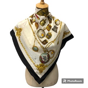 May include: A white silk scarf with a black border. The scarf features a repeating pattern of gold chains, pocket watches, and other antique trinkets. The text "La Fenice" is printed in the lower left corner.