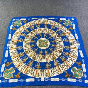 May include: A blue silk scarf with a gold and white pattern of chains and tassels. The center of the scarf features a circular design with a green and gold center.