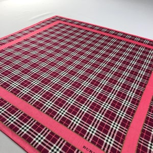 May include: A square scarf featuring a burgundy, black, white, and gold plaid design. It has a bright pink border. The word "BUR" is visible in the corner of the scarf.