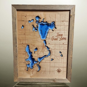 May include: A wooden framed wall art piece featuring a 3D topographic map of a lake. The lake is depicted in shades of blue, with the surrounding land in a light brown color. The text "Iowa Great Lakes" is visible.