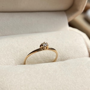 Puede incluir: Un anillo de oro con una pequeña piedra preciosa redonda y transparente engastada en una montura de seis garras. El aro es fino y pulido, con un acabado metálico liso. El anillo está colocado en posición vertical en una ranura de terciopelo.