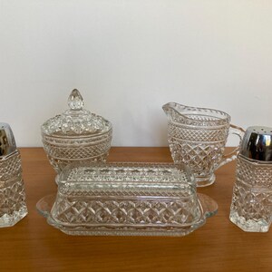 Puede incluir: Un juego de comedor de cristal transparente y vintage con un patrón de diamantes. El juego incluye un azucarero con tapa, una jarra de crema, una mantequera y dos saleros con tapas plateadas. Las piezas están dispuestas sobre una superficie de madera.