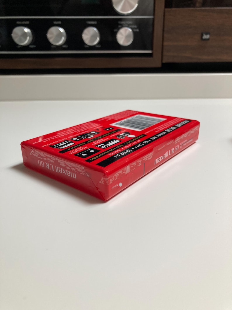 May include: A red Maxell UR 60 audio cassette tape in its original plastic shrink-wrap packaging. The rectangular box features white text, a barcode, and technical specifications including IEC TYPE I EQ 120 &micro;s. The branding Maxell UR 60 is visible.