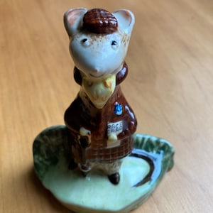Puede incluir: Una figura de cerámica de un ratón con un abrigo de cuadros marrón, un sombrero a juego y una bufanda amarilla. El ratón sostiene una tarjeta blanca con el texto RACE CARD impreso. La figura se apoya sobre una base verde con una cola pintada.