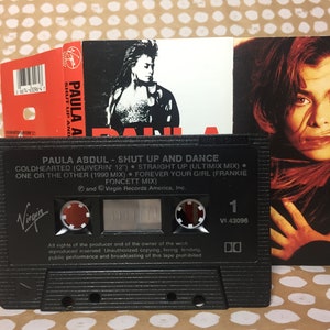 May include: A black cassette tape with a white label. The label has the text "PAULA ABDUL - SHUT UP AND DANCE" and lists the songs "COLDHEARTED (QUIVERIN 12")", "STRAIGHT UP (ULTIMIX MIX)", "ONE OR THE OTHER (1990 MIX)", "FOREVER YOUR GIRL (FRANKIE FONCETT MIX)". The label also has the Virgin Records logo.
