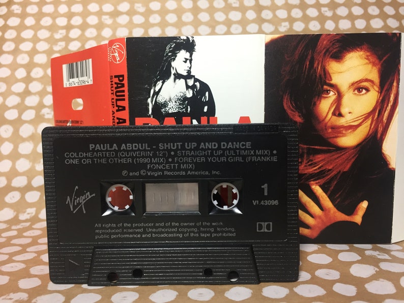May include: A black cassette tape with a white label. The label has the text "PAULA ABDUL - SHUT UP AND DANCE" and lists the songs "COLDHEARTED (QUIVERIN 12")", "STRAIGHT UP (ULTIMIX MIX)", "ONE OR THE OTHER (1990 MIX)", "FOREVER YOUR GIRL (FRANKIE FONCETT MIX)". The label also has the Virgin Records logo.