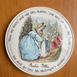 Puede incluir: Una placa de pared redonda de cerámica blanca con una ilustración de Beatrix Potter de la Sra. Coneja y sus hijos. El texto dice: 'Now, my dears,' said old Mrs. Rabbit, 'you may go into the fields......... but don't go into Mr. McGregor's garden.'
