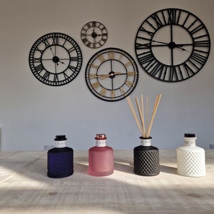 Puede incluir: Cuatro botellas de vidrio decorativas en vidrio negro, blanco, rosa y azul. Las botellas tienen un diseño texturizado y están llenas de aceite perfumado. Las botellas están dispuestas sobre una mesa de madera frente a una pared blanca con cuatro relojes negros y dorados.