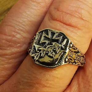 May include: A silver-toned metal ring featuring an embossed Iron Cross symbol and a laurel branch design on the face. The band has intricate textured detailing along the sides with the text "FUR BEILAGE" engraved on the right side of the ring.