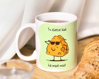 May include: White ceramic mug with a green background and a cartoon illustration of a smiling piece of cheese wearing sunglasses. The text "Tu cheese badi hai mast mast" is printed on the mug.