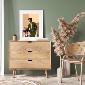 May include: A framed illustration of a man in a green suit standing in front of a balcony with a glass in his hand. The illustration is on a wooden dresser with three drawers. A wicker chair and a dried flower arrangement are in the foreground.