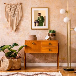 May include: A mid-century modern wooden dresser with three drawers and gold hardware. The dresser is decorated with a framed print of a man in a green suit holding a glass of wine. A potted plant and a lamp with three white globes are on the dresser.