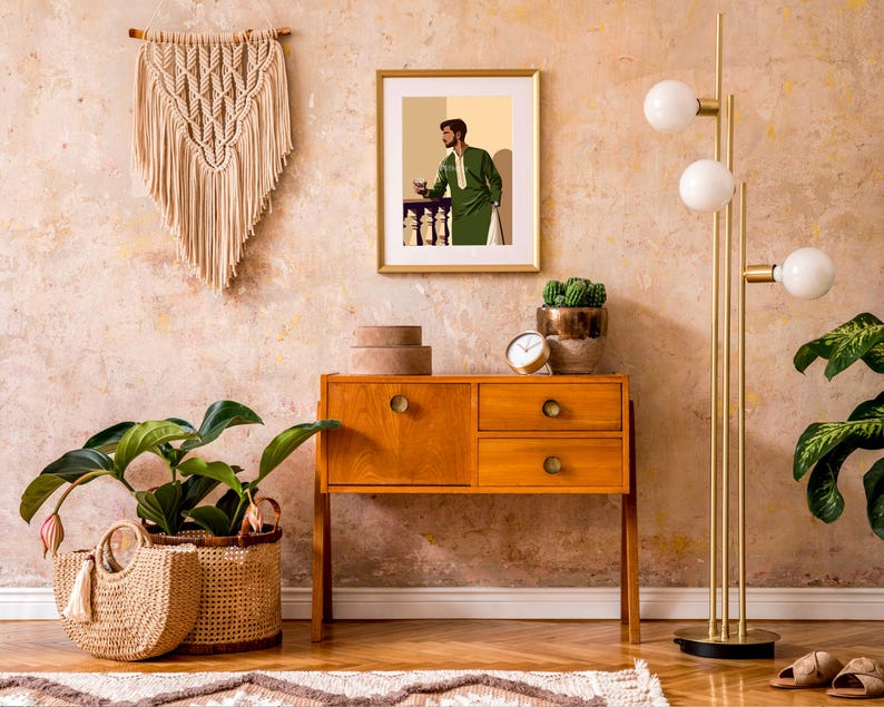 May include: A mid-century modern wooden dresser with three drawers and gold hardware. The dresser is decorated with a framed print of a man in a green suit holding a glass of wine. A potted plant and a lamp with three white globes are on the dresser.
