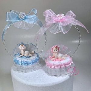 Puede incluir: Dos figuritas de bebés, una azul y otra rosa, están sentadas en un adorno para pastel. Las figuritas están en un pastel blanco con una cinta azul y rosa alrededor de la base. El adorno para pastel está hecho de plástico transparente y tiene un lazo en la parte superior.